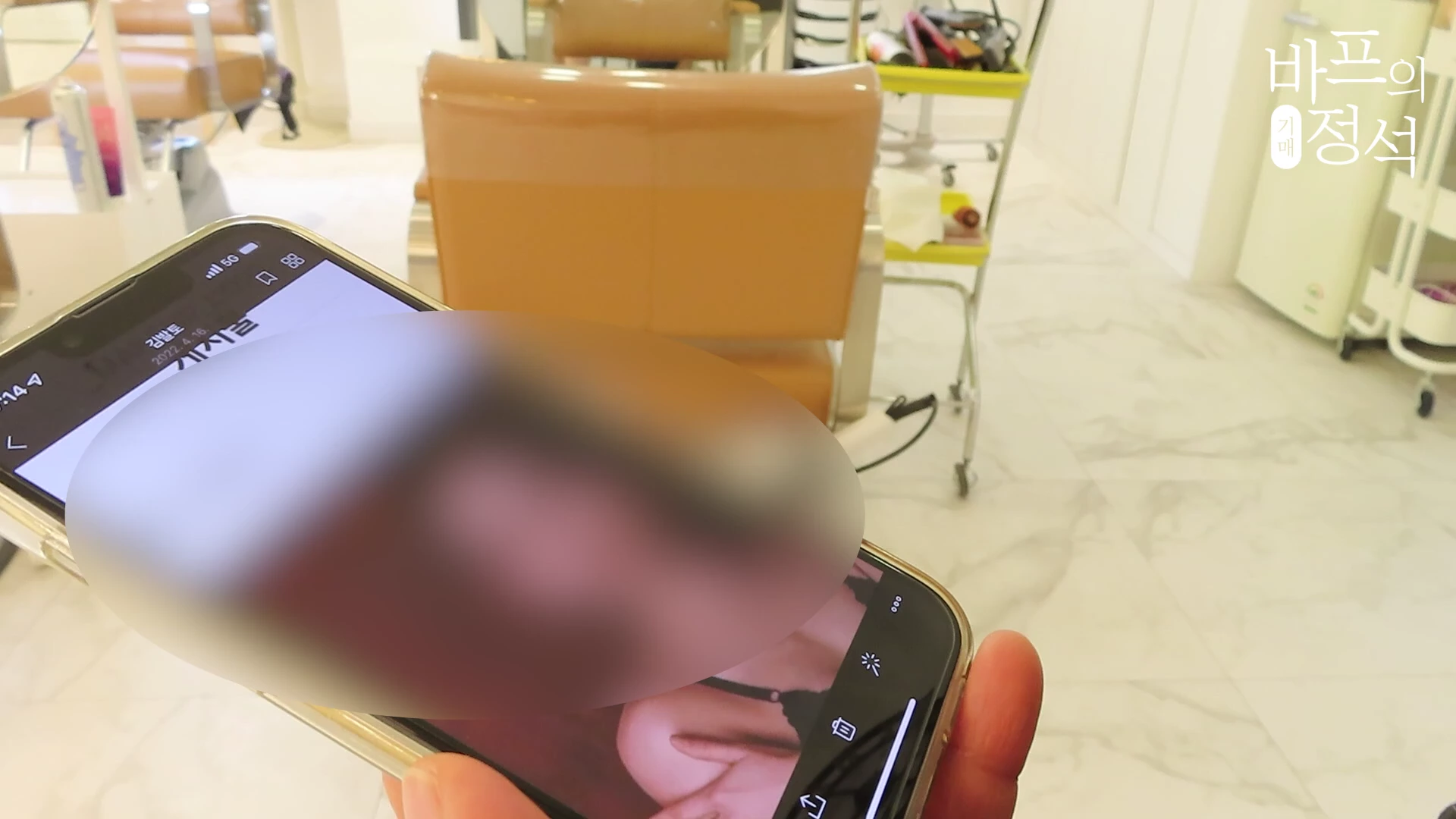 미용실을 배경으로 한 손이 들고 있는 스마트폰 화면에 메이크업과 헤어 스타일 참고용 사진이 떠 있다.