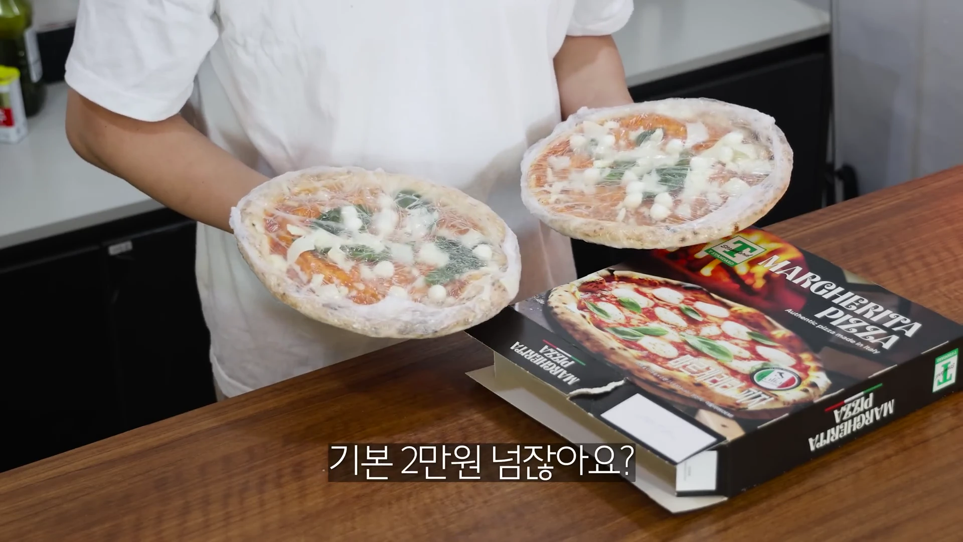 흰 티셔츠를 입은 사람이 비닐로 개별 포장된 두 개의 냉동 마르게리따 피자를 양손으로 들고 있고, 그 앞에는 빈 피자 상자가 놓여 있다.