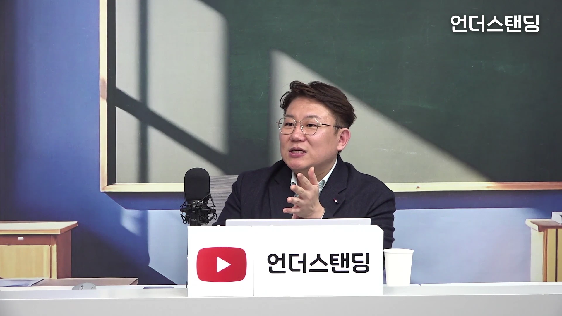 안경을 쓴 남성이 스튜디오 책상 앞에 앉아 손짓하며 대화하고 있는 모습. 뒤로는 칠판 배경이 보이며 책상 위에는 마이크와 채널 로고가 적힌 흰색 표지판이 놓여 있다.
