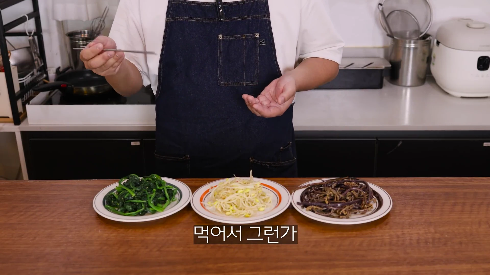 앞치마를 두른 사람이 주방 조리대 앞에서 시금치, 콩나물, 고사리 나물 세 접시를 소개하고 있다.