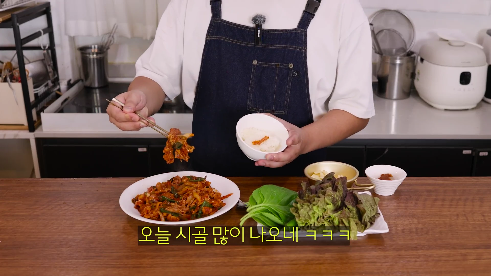 앞치마를 두른 요리사가 한 손에는 밥그릇, 다른 손에는 젓가락으로 제육볶음을 집어 든 채 식탁 앞에 서 있다. 앞쪽에는 제육볶음 접시와 쌈 채소가 놓여 있고 화면 하단에는 자막이 달려 있다.