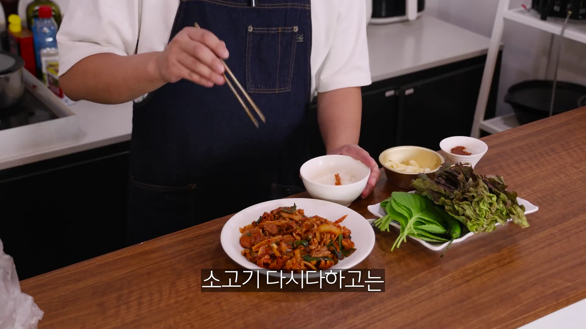 앞치마를 두른 사람이 젓가락을 들고 완성된 제육볶음 접시 앞에 서 있는 모습. 식탁 위에는 쌈 채소와 작은 공기들이 놓여 있다.