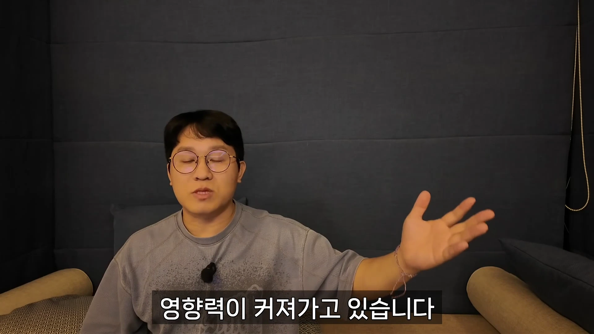 안경을 쓴 남성이 어두운 배경 앞에서 이야기를 하며 한쪽 손을 들어 올리고 있는 모습의 영상 캡처 화면