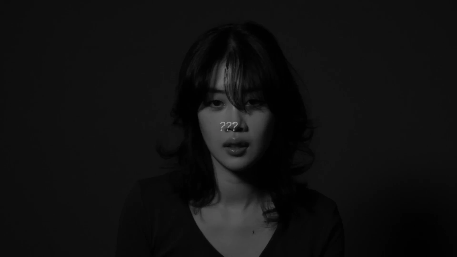 어두운 배경 속에서 카메라를 정면으로 응시하는 여성의 얼굴 위로 물음표 세 개가 겹쳐져 있는 흑백 영상 캡처 화면입니다.