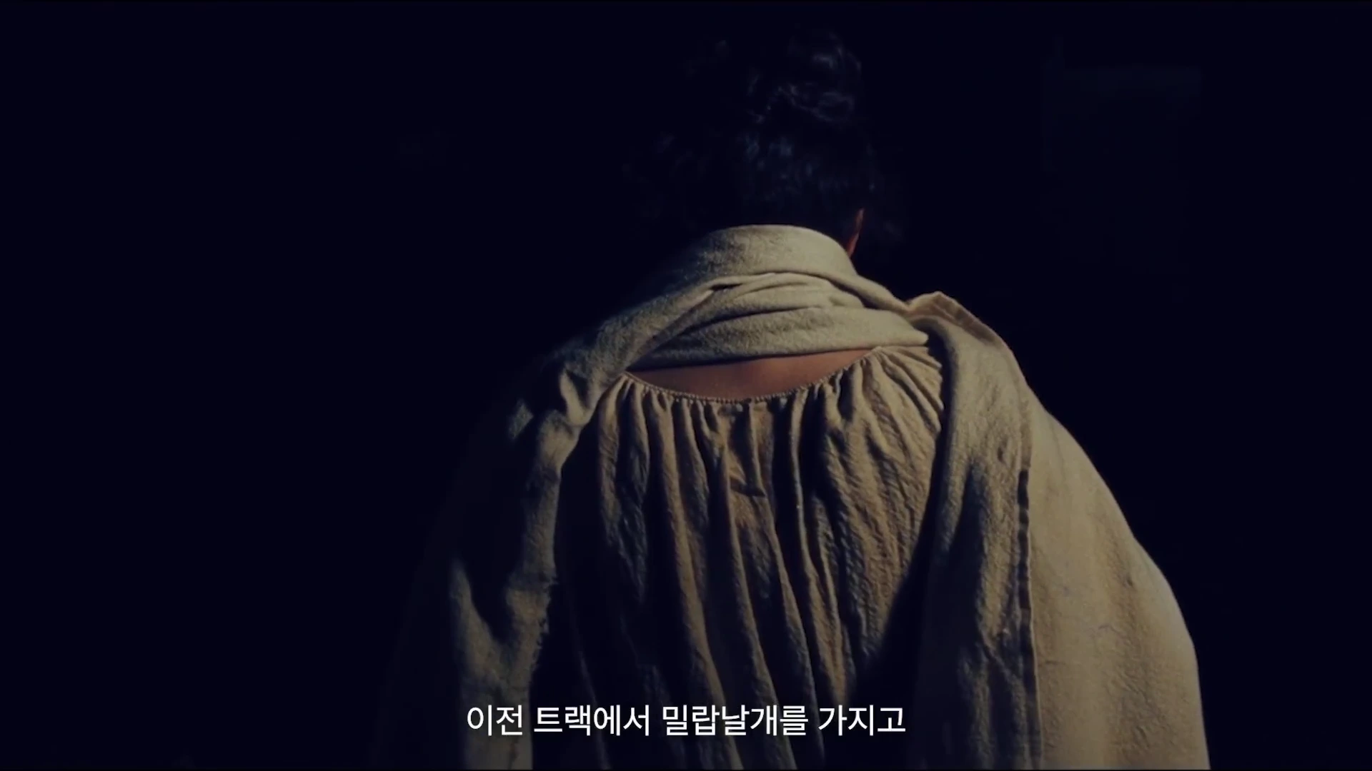 어두운 배경 속에서 베이지색 숄을 걸친 인물의 뒷모습이 부드러운 조명을 받으며 서 있고, 하단에는 한국어 자막이 새겨져 있습니다.