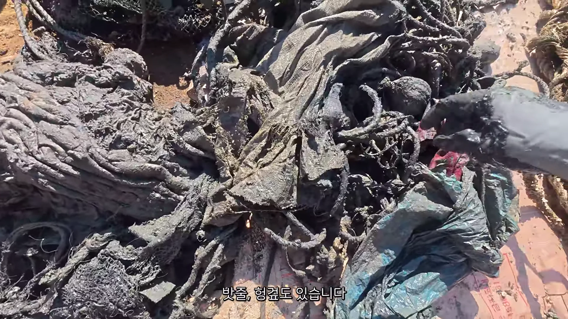바닷속 진흙과 뒤섞여 형체를 알아보기 힘들 정도로 엉킨 천 조각, 비닐, 밧줄 등 해양 침적 쓰레기가 쌓여 있고, 검은 장갑을 낀 손이 이를 들추고 있는 모습.