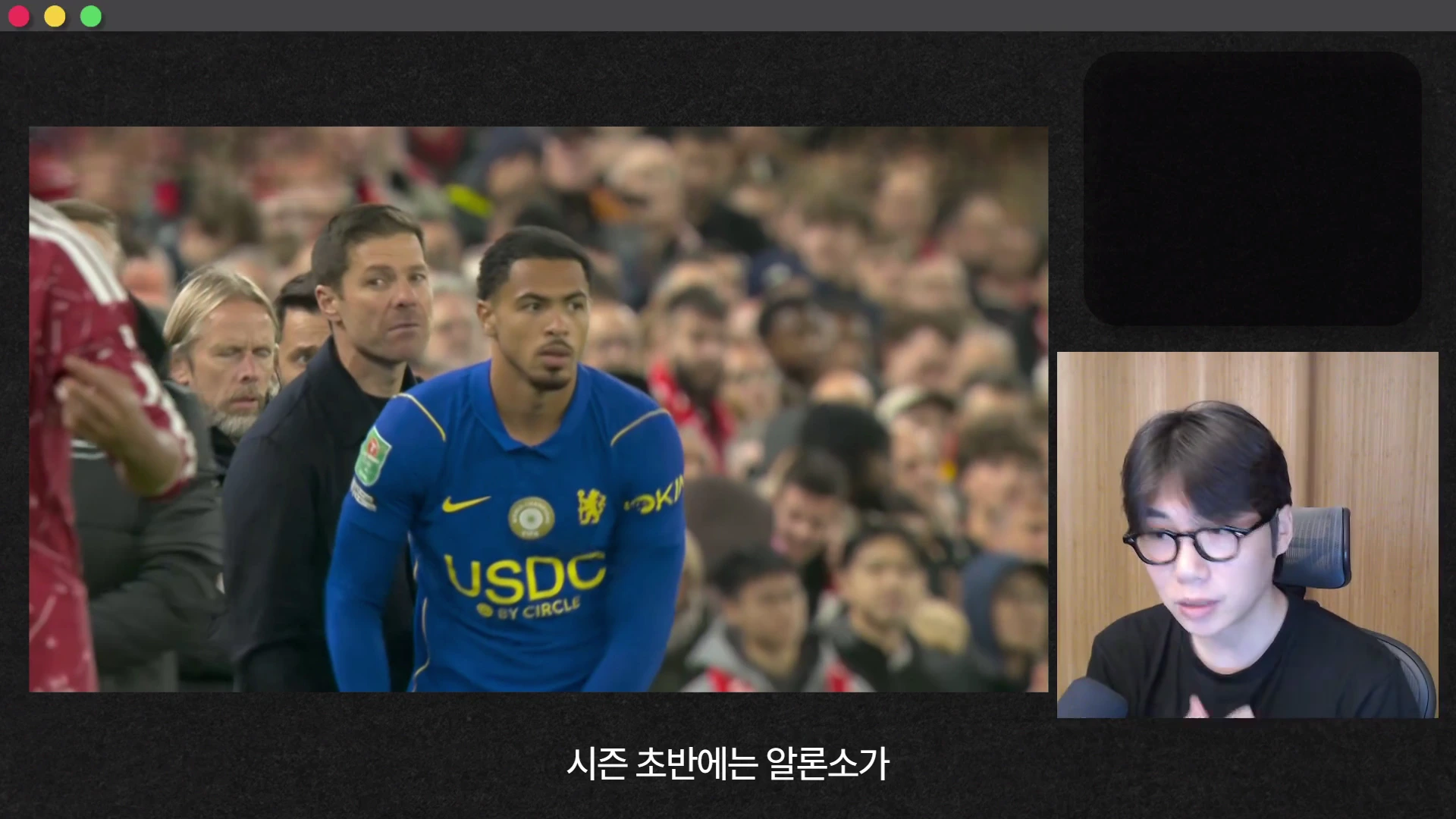 축구 경기장에서 파란색 유니폼을 입은 선수와 감독이 경기를 지켜보고 있으며, 화면 우측 하단에는 안경을 쓴 남성이 마이크 앞에서 이야기하는 모습이 담긴 유튜브 영상 캡처 화면입니다.