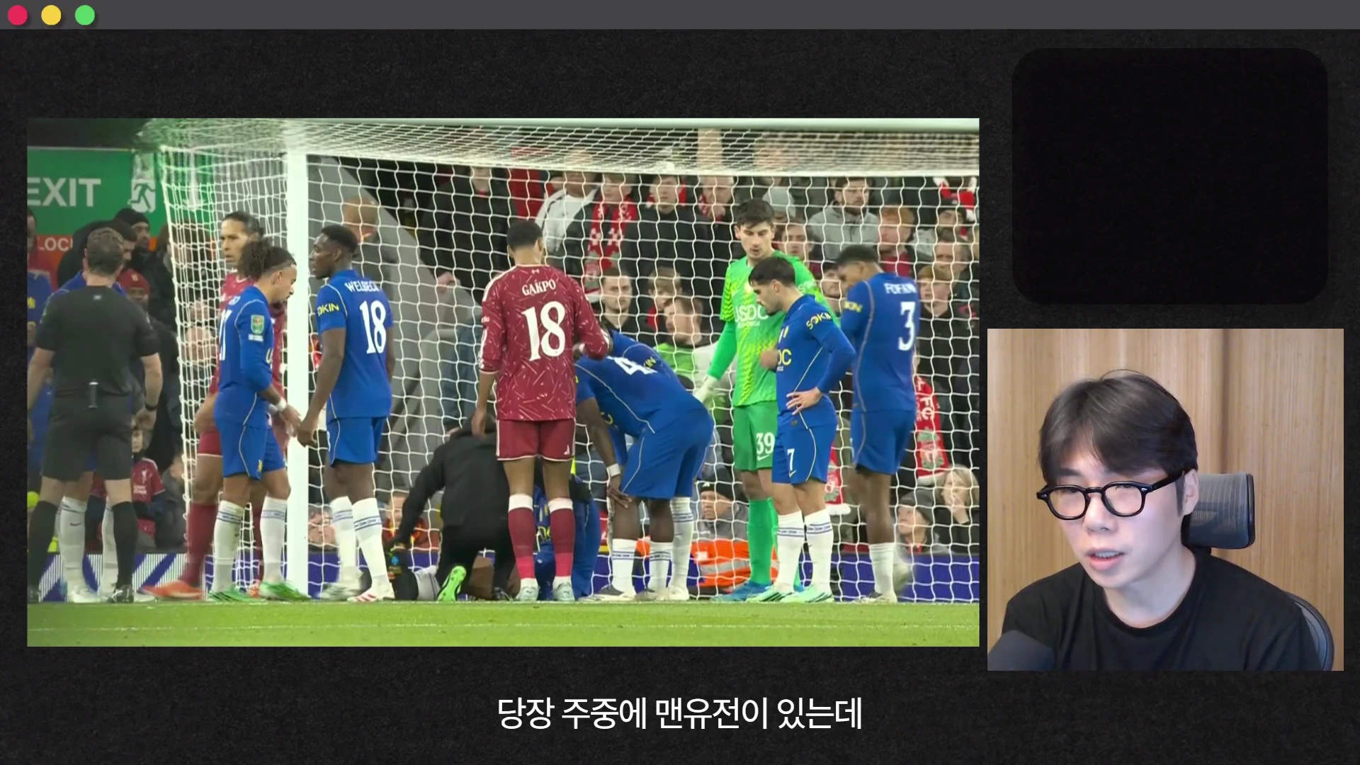 축구 경기 중 그라운드 위에서 선수들이 부상당한 동료를 살피는 모습이 왼쪽에 보이고 오른쪽 하단에는 안경을 쓴 남성이 화면을 바라보며 이야기하고 있다.