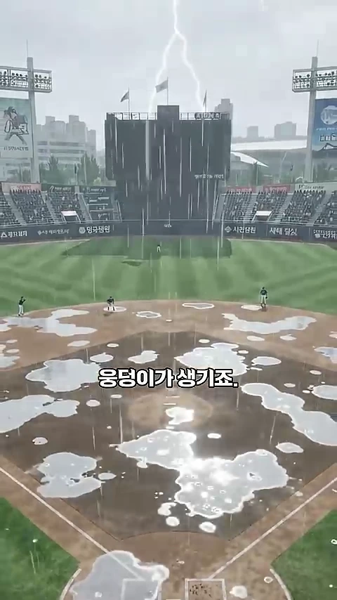 비 내리는 야구장의 내야 흙바닥에 크고 작은 물웅덩이가 여러 개 고여 있는 모습이 담긴 영상 캡처 화면이며, 배경에는 전광판과 관중석이 보이고 화면 중앙에 '웅덩이가 생기죠'라는 자막이 적혀 있다.