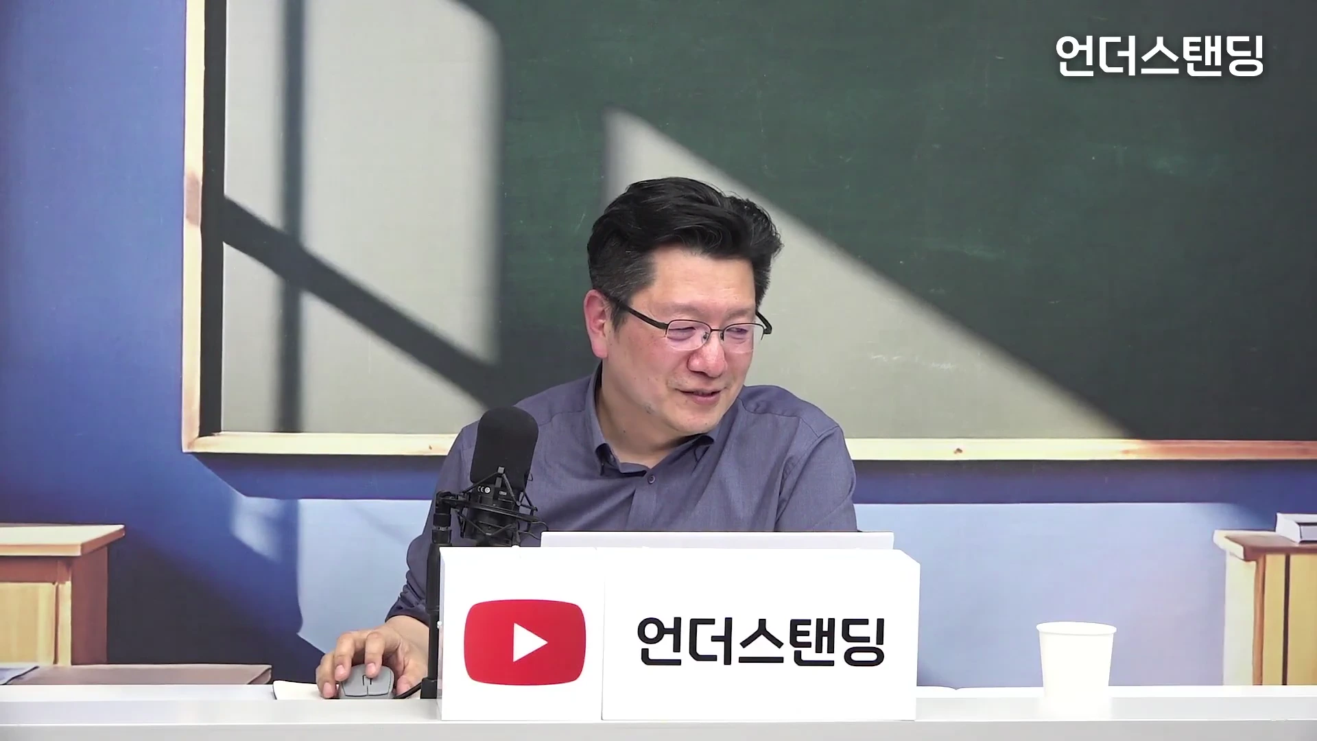 안경을 쓴 중년 남성이 스튜디오 책상에 앉아 마이크 앞에 대고 이야기하고 있는 모습